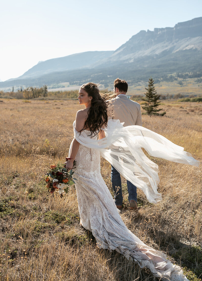 A bride and groom walk through a golden Montana field surrounded by mountains, her lace dress and veil flowing in the wind, captured by Sydney Breann Photography during their intimate elopement.