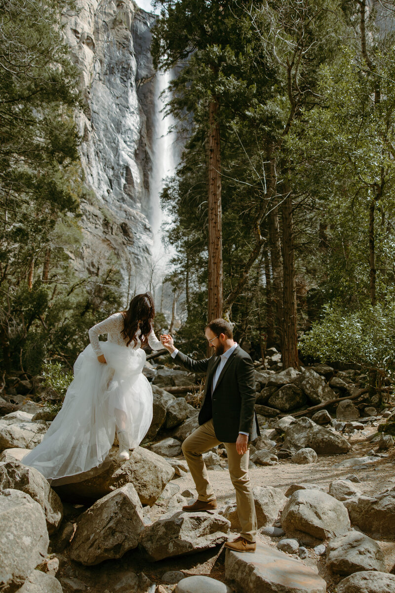Bride walking down off a rock and her groom helping her with a beautiful waterfall in the background