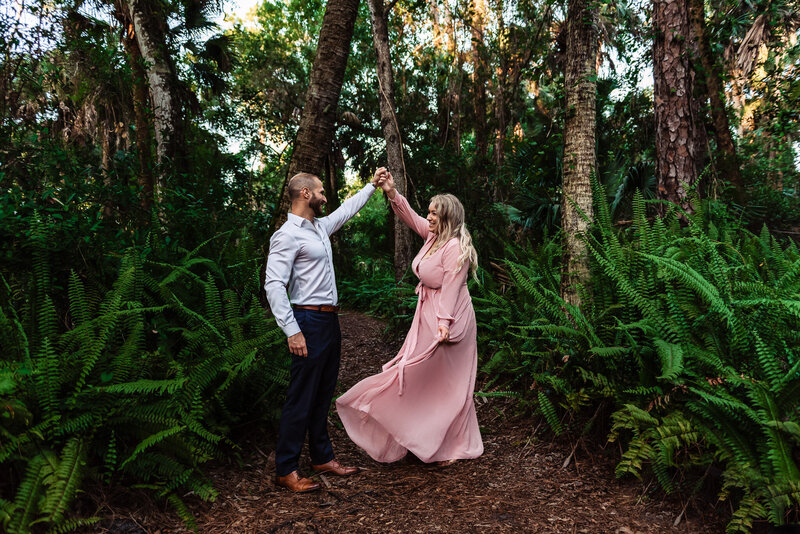 A couple dancing in ferns. Man is wearing clue pants and white shirt and girl in a flowing pink dress being spun.