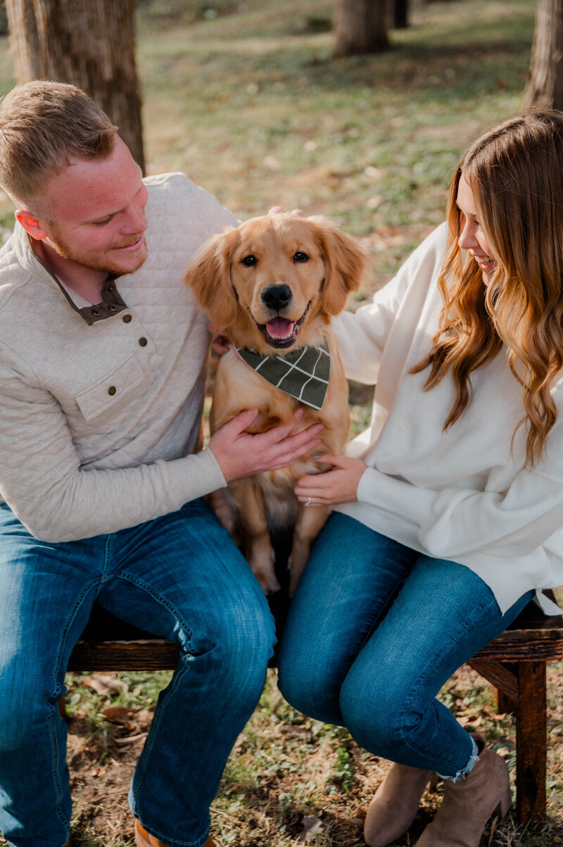 A couple poses smiling at their dog and their dog smiles at the camera.