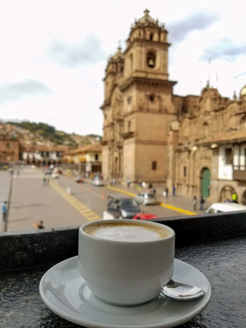 A frothy cappuccino on a balcony overlooking Plaza de Armas in Cusco, Peru, with the historic stone façade of the Church of the Society of Jesus in the background ©Stephanie Dosch | theViatrix Bespoke Peru Travel Itinerary