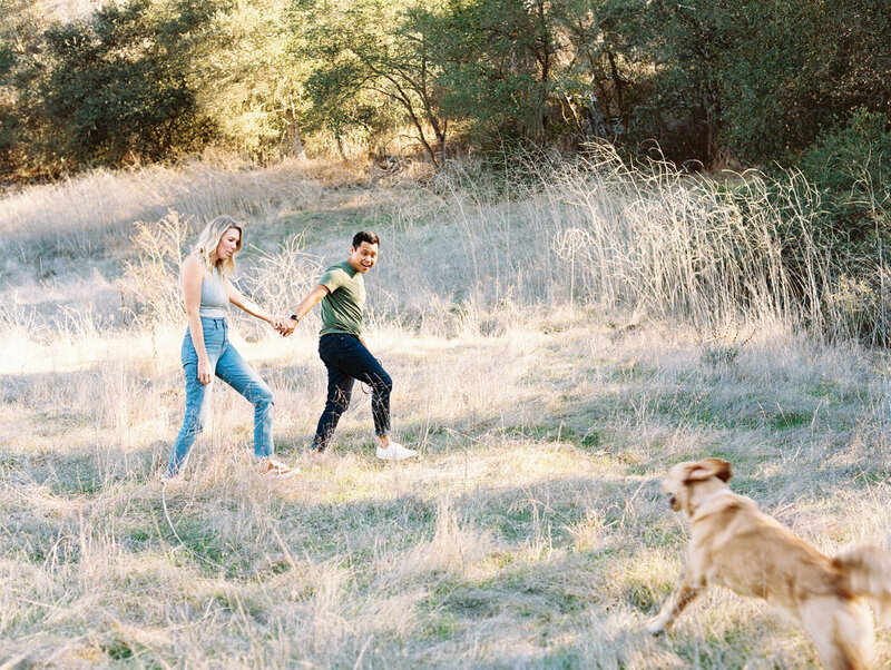 fiances holding hands walking while their dog sprints towards them and is blurry from how fast