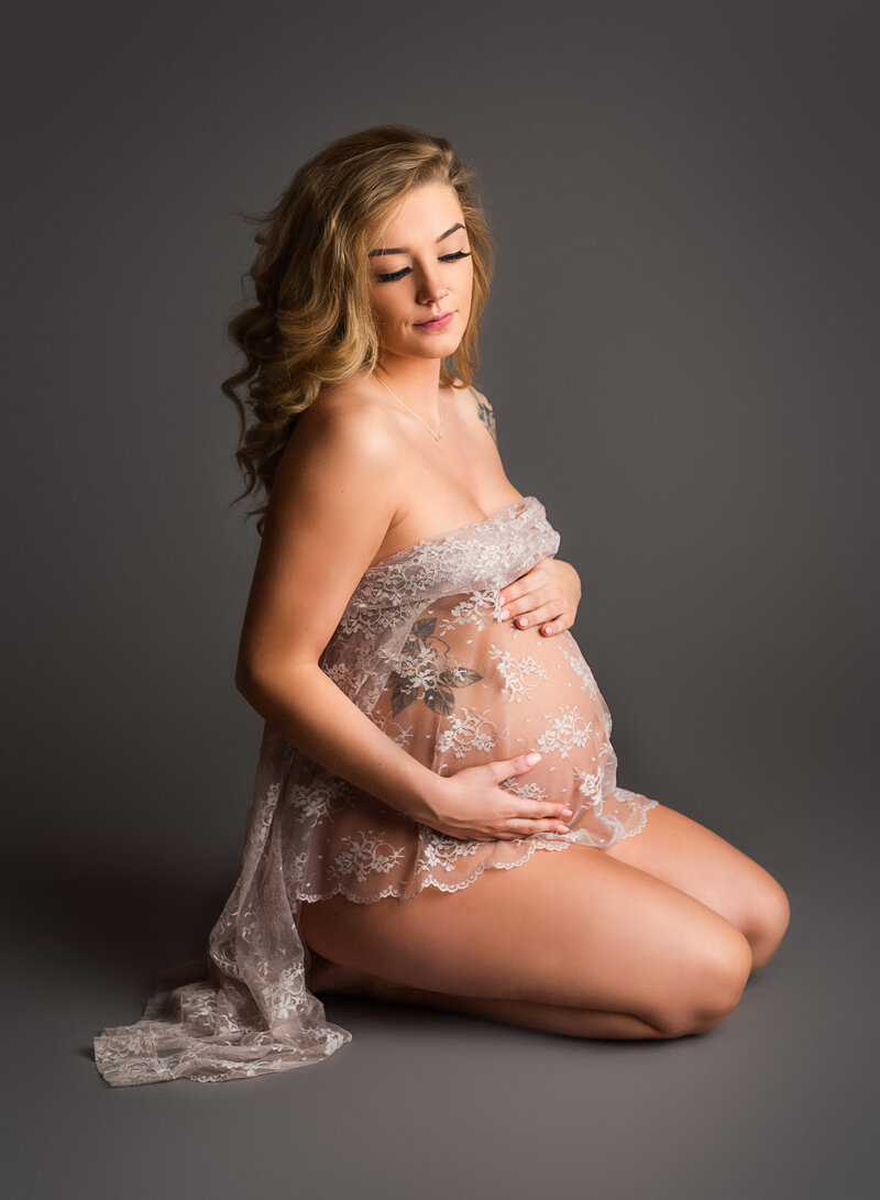 Pregnant woman kneeling, draped in a sheer lace fabric, gently holding her belly