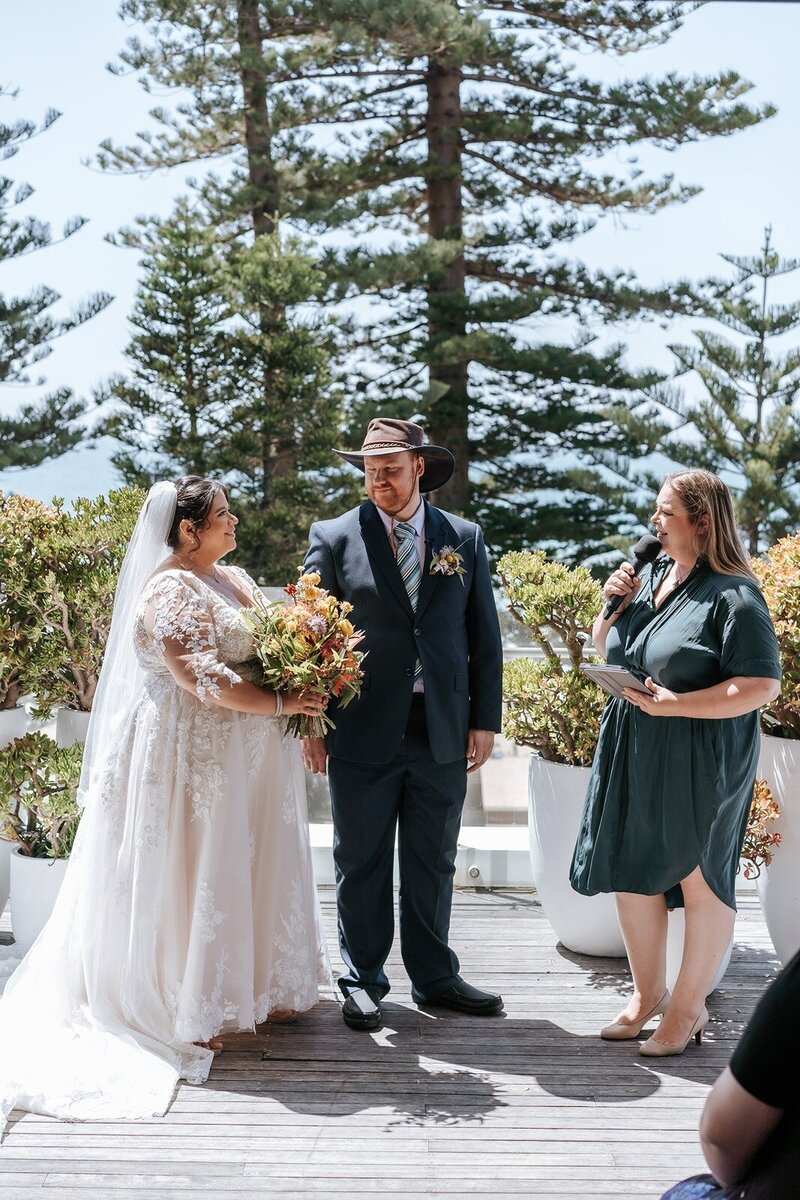 A bride in a lace dress holding a bouquet standing next to her groom and the celebrant