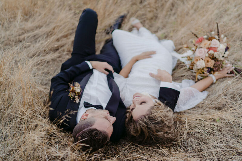 Bride and groom laying in dried grass field.