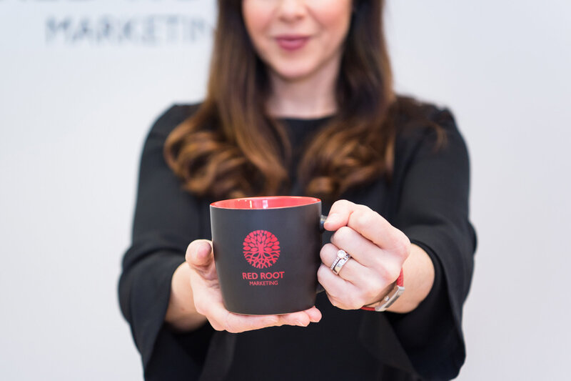 A woman holding a black mug with a Red Root Marketing logo.