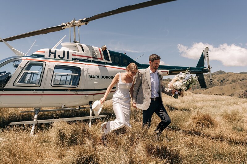 a bride and groom climb out of a helicopter in blenheim on a golden grass hilltop on thier wedding day