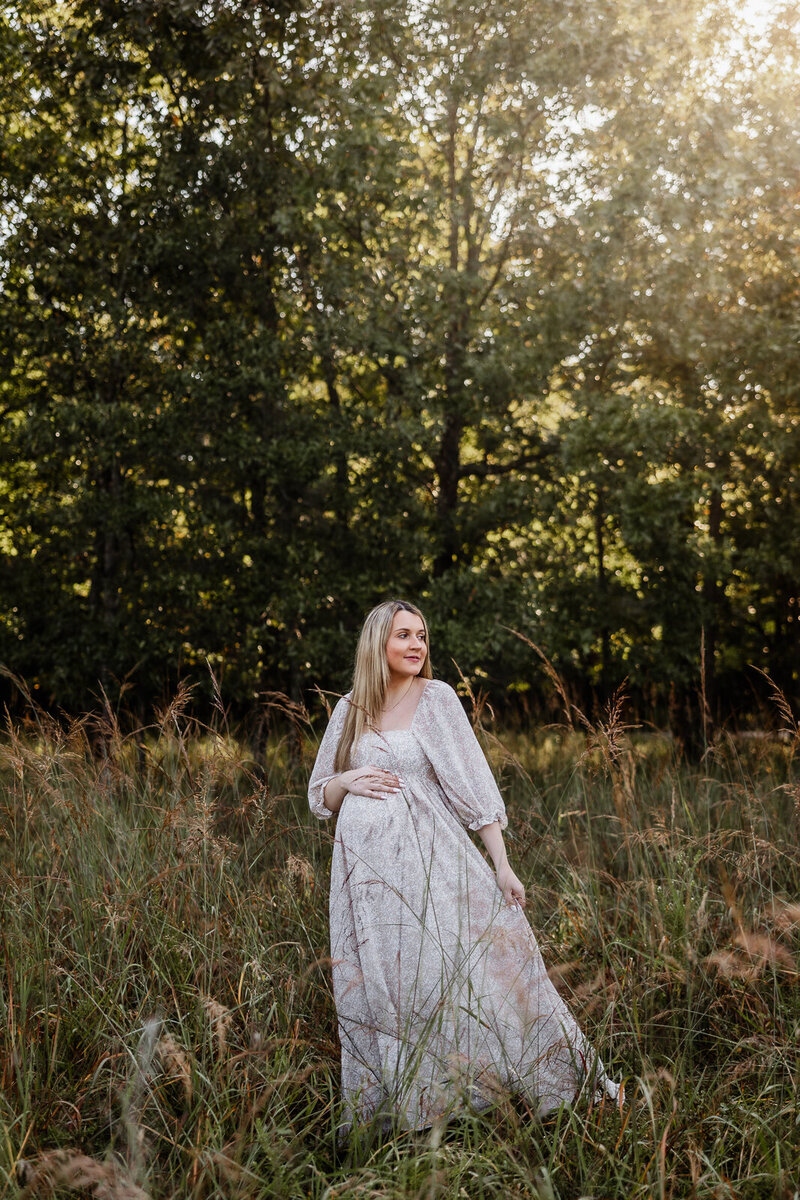 Maternity photo of mom-to-be standing in a meadow with sunlight streaming behind her.