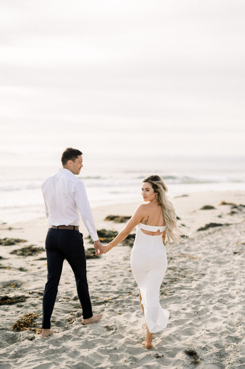 Karla & Seath - San Diego, California - Couple walking on beach in white outfits at sunset.