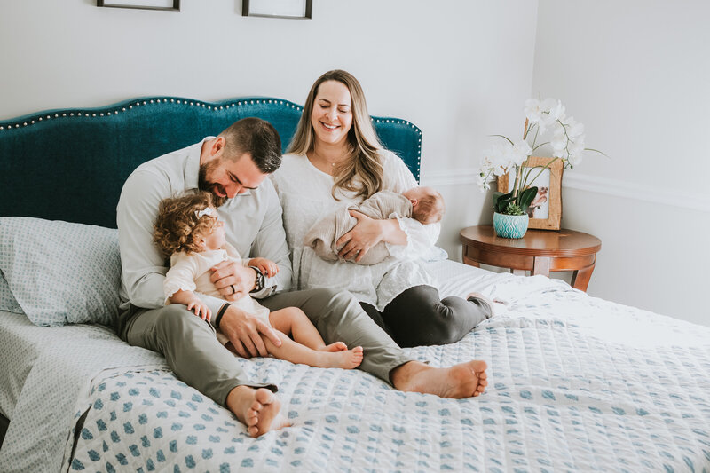 Mom, dad, and sibling sitting on a bed smiling and laughing while mom is holding baby girl.