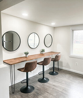 A modern dining area features a wooden counter accompanied by three brown bar stools and adorned with two small potted plants. Three round mirrors reflect the light-colored walls, while a window on the right adds openness to this tastefully designed space. Explore more at https://halcyon-watson-1.showit.site/the-venue.