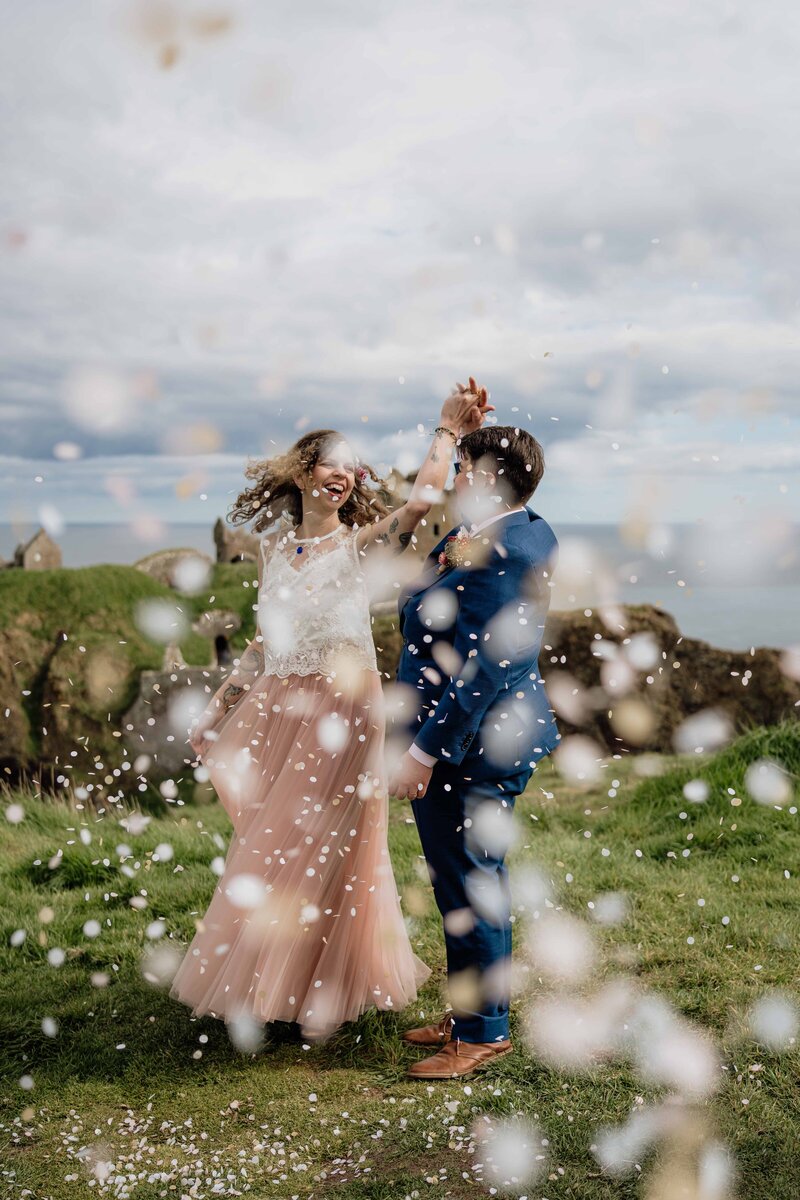 An LGBTQ+ couple stand on the side of a cliff and one twirls another as confetti falls all around them. Dunnottar Castle and the Aberdeenshire coast can be seen behind them as they’re photographed. 