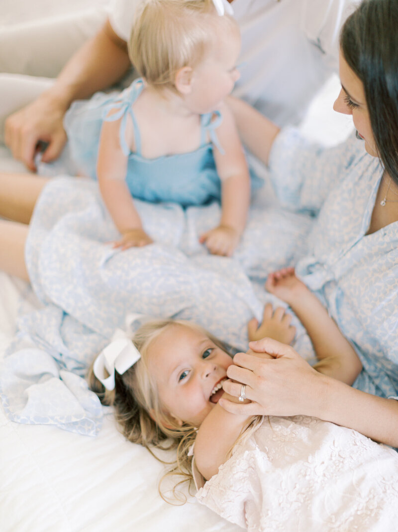 Blonde girl lays laughing in mothers lap, who is wearing a blue dress, while also holding her and her sister, also in blue, inside Little Rock studio, taken by Bailey Feeler Photography