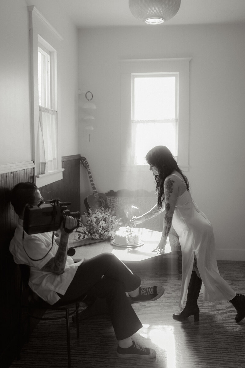 Black and white wedding photography of a bride lighting candles on a cake while the groom films with a vintage camcorder. Editorial and cinematic Greenville wedding photography by Dani Lacey Photographs, capturing artistic and authentic South Carolina wedding moments.