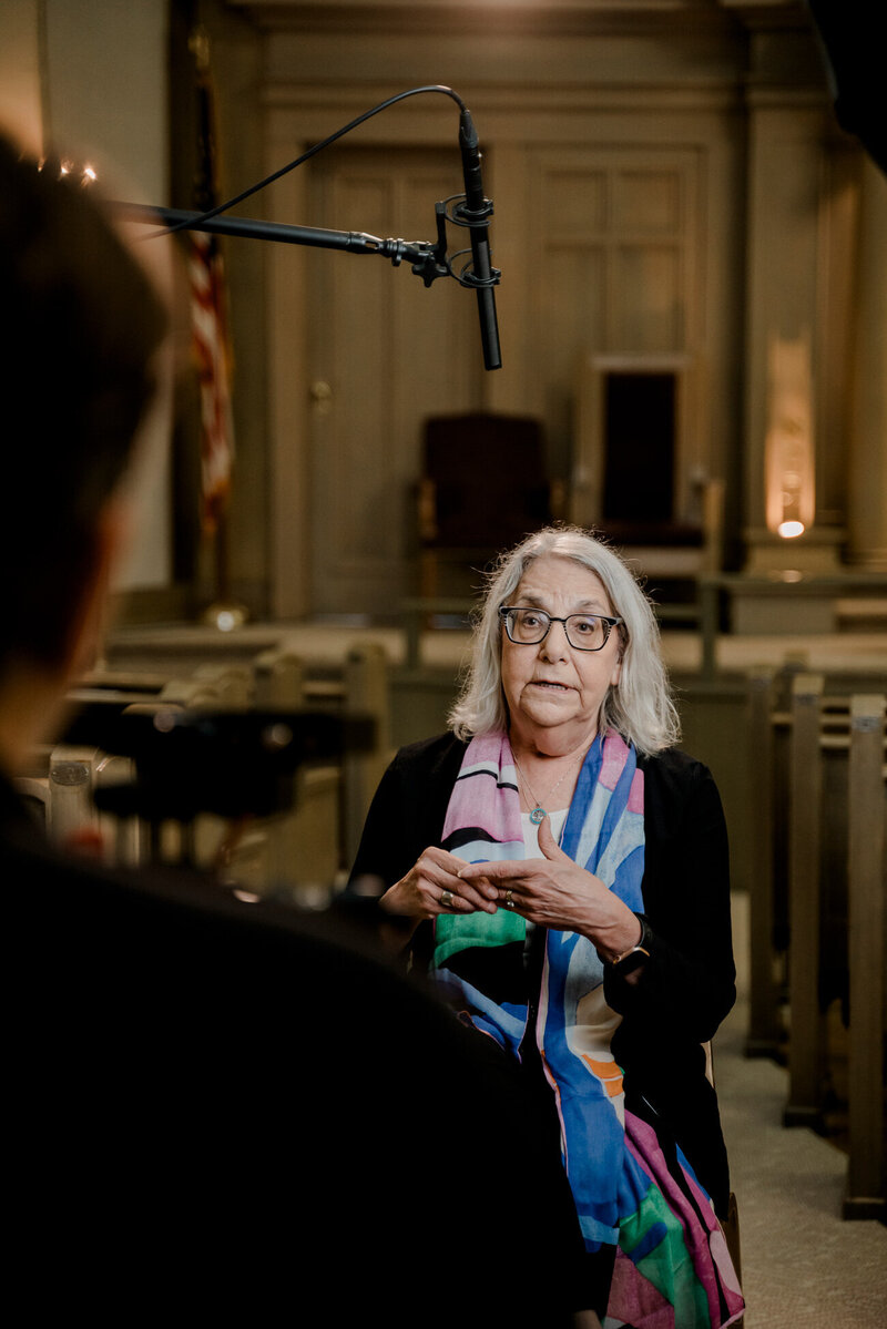 a woman with grey hair and a bright  colorful shall sits under lights and a boom microphone  with a video producer in the foreground