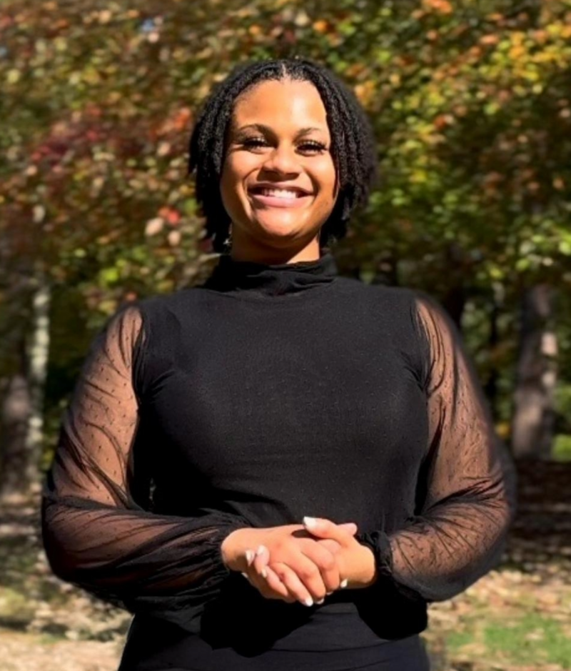 Angel is a young black woman with dark ringlet curls that go to her jaw, dark brown eyes, with a full beautiful smile and warm skin. She is wearing a black long sleeved blouse.