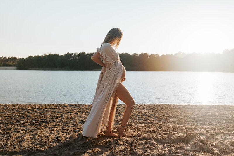 Zwangere vrouw in een beige jurk poseert aan 't zand bij zonsondergang tijdens zwangerschapsshoot in Alphen.
