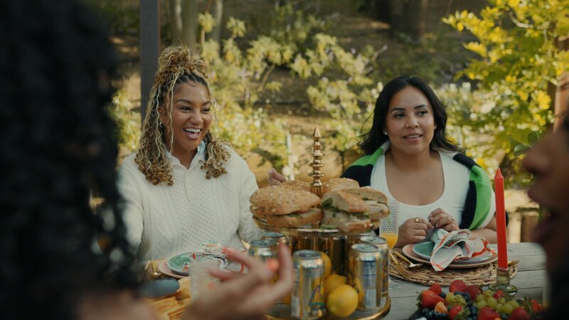 Two women sat at a brunch table laughing, with bagels and canned drinks.