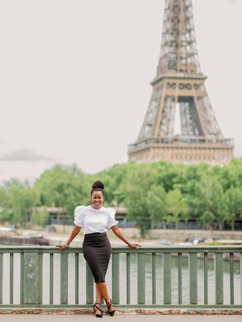 a black woman with a white top and black skirt standing in front of the eiffel tower on a bridge in Paris