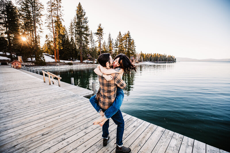A couple embraces joyfully on a wooden dock at Lake Tahoe, with the sun setting behind the snow-dusted pine trees and calm waters reflecting the golden light.