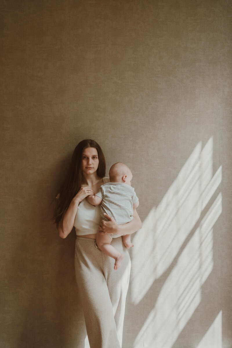 Een foto van een moeder met een baby tegen een muur aan terwijl de moeder recht in de camera kijkt