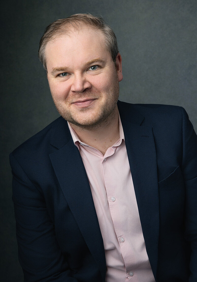 Headshot of a man taken in studio for a finance company He is wearing a dark suit and pink shirt without a tie and smiling at the camera.