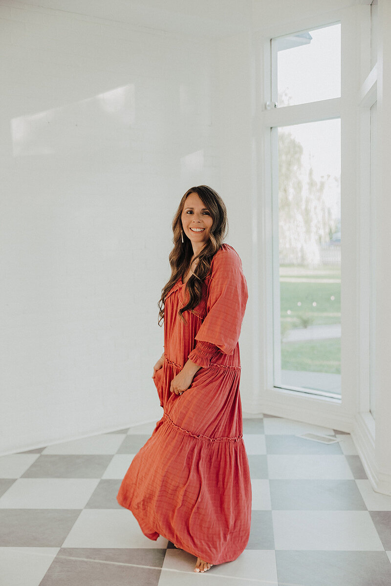 Patty in an orange dress against a white wall with a window 