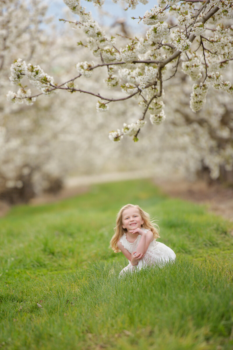 Little girl wearing a white dressing sitting the grass in an orchard in Caldwell, Idaho during photography session with Boise photographer Tiffany Hix