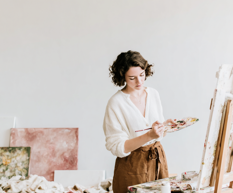 Woman painting in a sunlit studio surrounded by abstract canvases, representing the creative transformation and artistic empowerment of clients in Jasmina Kimova’s brand reinvention experiences