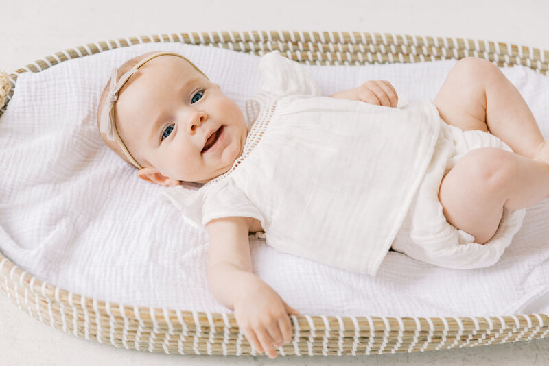 An infant girl smiles while laying in a woven basket in a white shirt and headband