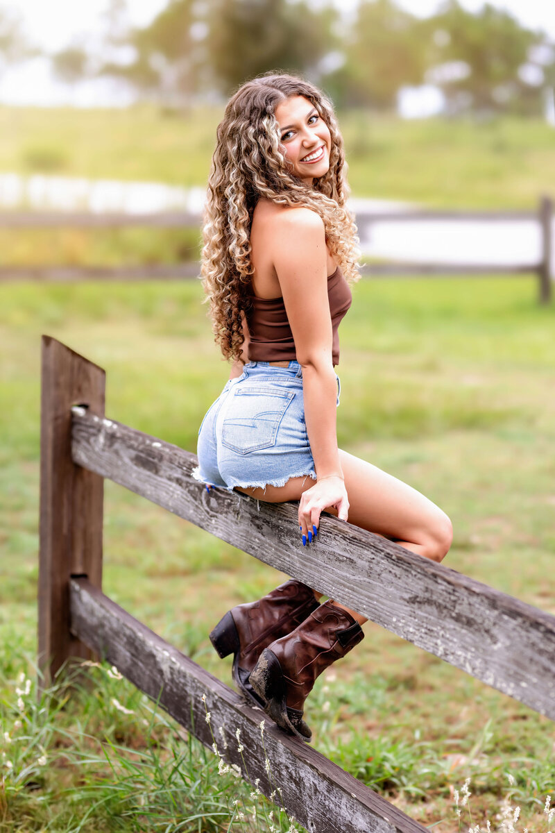 High school senior in jean shorts and brown top smiling in a golden field during a senior session in Winter Garden, Florida.
