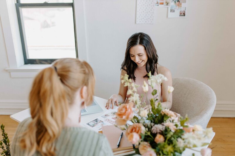 2 women sitting across at a desk, one with dark hair and other with red,blonde hair, flowers and paperwork and laptop on the desk