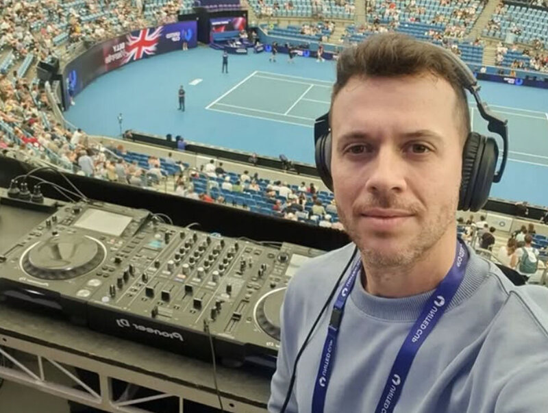 DJ wearing headphones taking a selfie with Pioneer DJ equipment and a live United Cup tennis match in the background