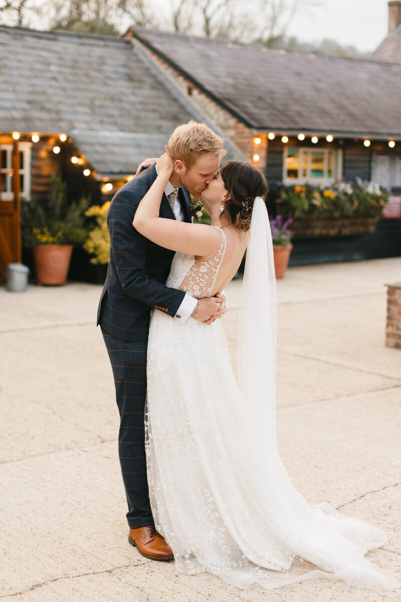 A newlywed couple embraces and kisses outdoors at Upwaltham Barns. The bride wears a white, lace wedding dress with a long veil, and the groom is in a dark checkered suit. They are surrounded by rustic buildings adorned with string lights, creating a magical ambiance.