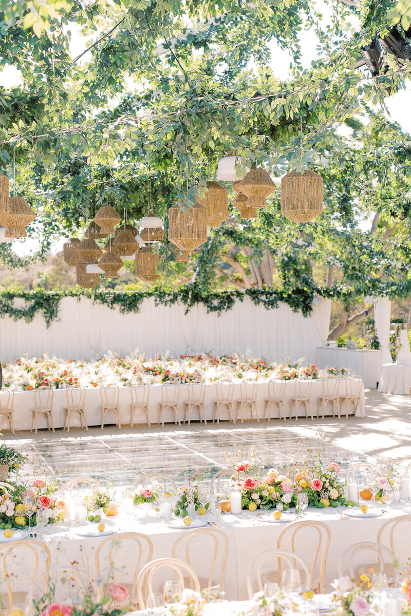 Outdoor wedding reception space, and glass dance floor. The white tables are lined with yellow and pink flowers, and there is greenery and wicker baskets hanging from the wire tent above.