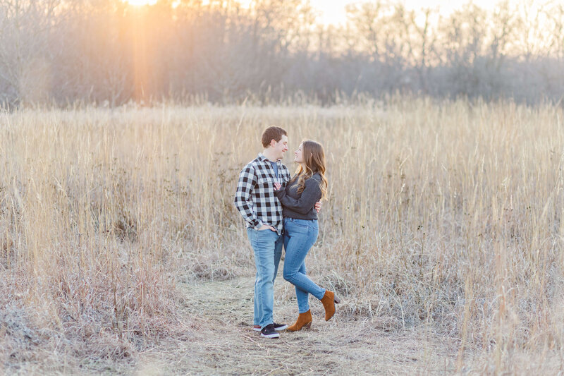 A man and woman stand in an open field as the sun sets smiling at each other.