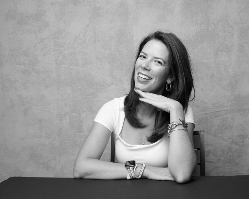 Black and white portrait of a woman sitting at a table, playfully posing with her chin resting on her hand and a bright, confident smile. She wears a fitted white top, hoop earrings, and stacked bracelets, exuding warmth and joy. A lively, unfiltered moment from 'THIS IS ME – A Photography Experience by Katie Campbell' in Atlanta, GA, celebrating personality and self-expression.