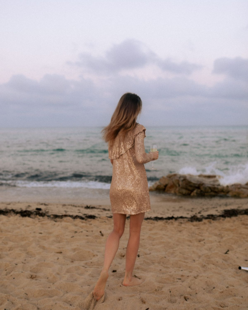 Woman in a sparkly dress walking along the beach during golden hour, facing the ocean