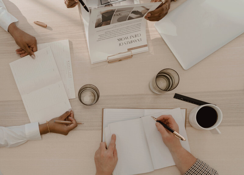 “Collaborative workspace meeting at Sable & Rose—hands of diverse professionals writing in planners and reviewing client work documents on a light wood desk. Essentials like a laptop, coffee, water glasses, and stationery reflect a modern, organized approach to strategic PR and brand strategy and development.