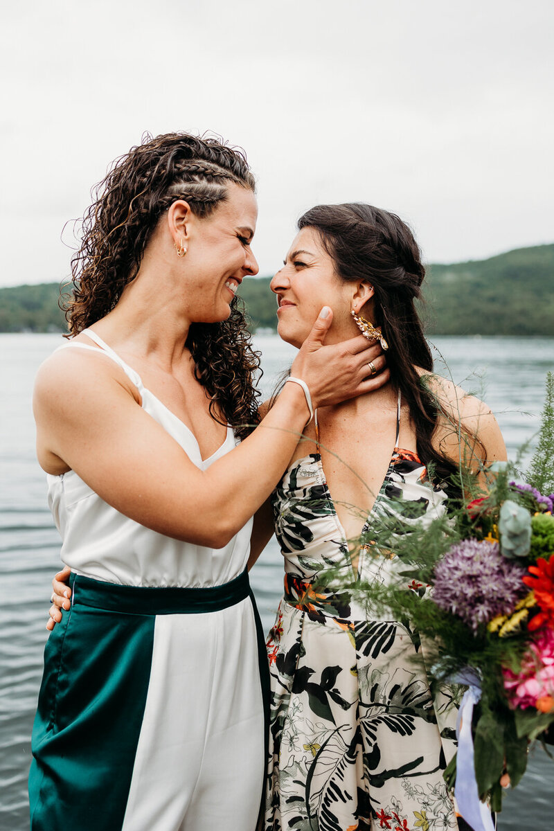 Newlywed brides embrace and lean in for a happy kiss by the water captured by their LGBTQ+ Saratoga Springs wedding photographer