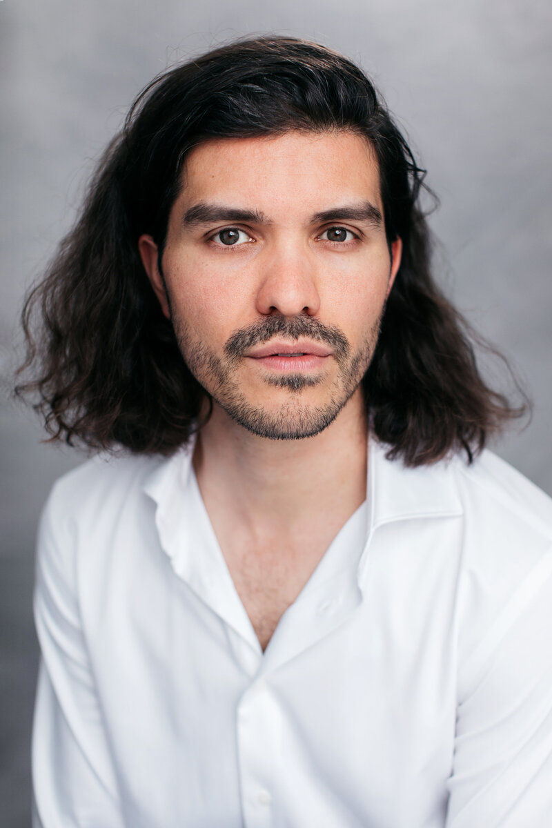 Casting headshot van man met lang donker haar in wit overhemd voor grijze achtergrond.