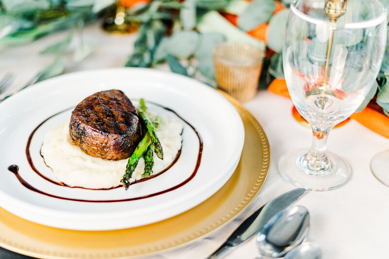 Photograph of a steak delicately plated for serving with mashed potatoes and asparagus on a tablescape set with fresh greenery and taper candles.