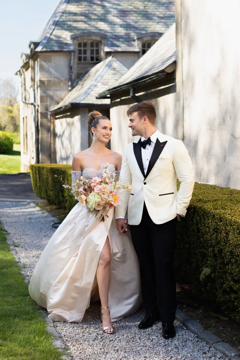 Wedding Photographers in RI | Kelsey Sheehan Photography Timeless Rhode Island Weddings | A bride and groom stand on a garden path, smiling at each other. She holds a bouquet and wears a white off-shoulder gown; he wears a white tuxedo jacket.