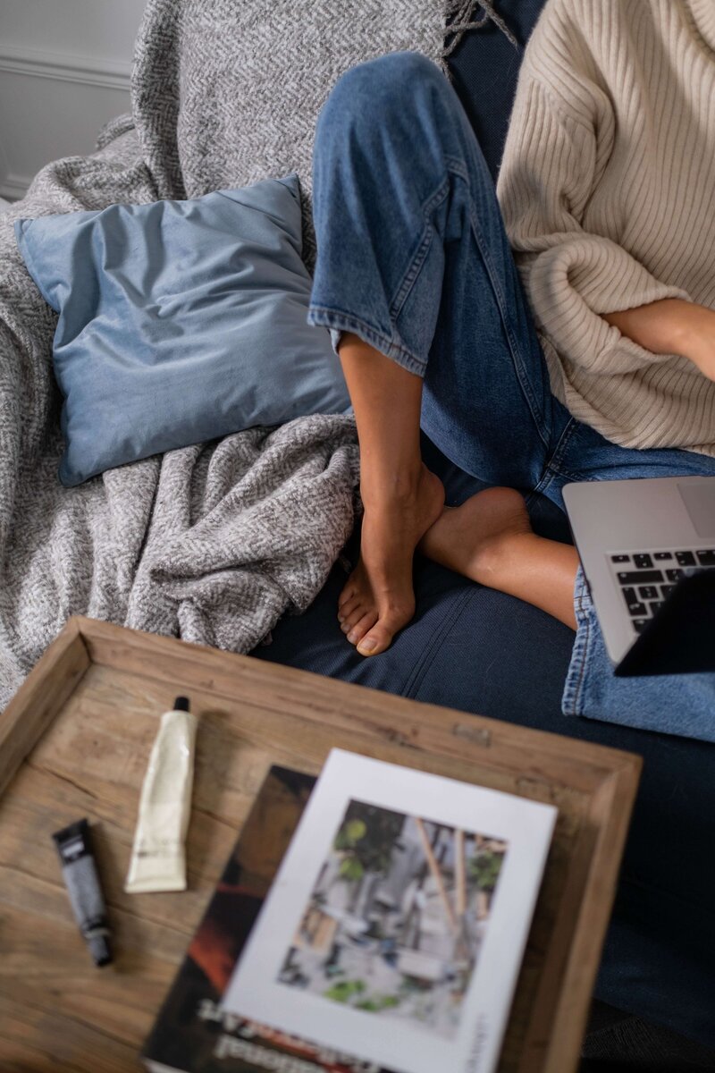 Cozy minimal scene of a woman's lower half on a blue couch with some lifestyle items like a blanket pillow book. She works on a laptop.