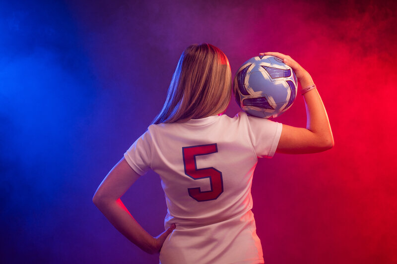 A person in a soccer jersey facing away and holding a soccer ball up in their bicep
