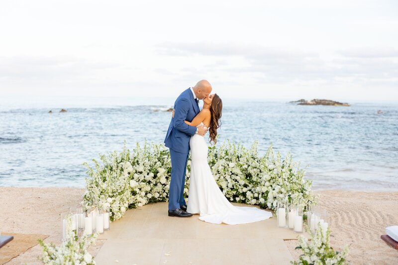 Newlywed couple, Nick and Brooke, embrace and kiss on a beach with the ocean in the background while standing amongst a half-ring of flowers and candles