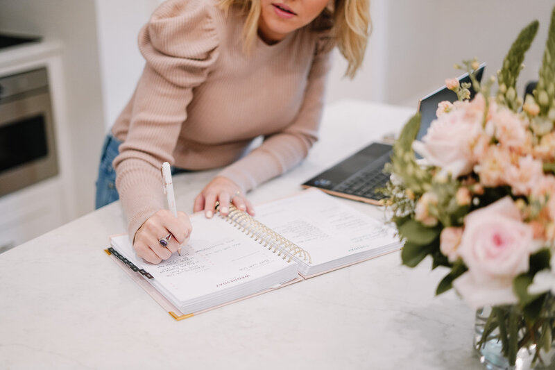 Woman in soft taupe sweater at white marble desk with laptop writes in her planner