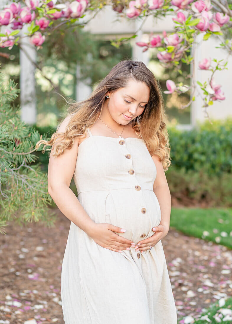 A pregnant woman wearing a cream colored sundress holds her belly while she stands in front of blossoming magnolia trees on the grounds of BYU in the spring. Photo taken by Utah maternity photographer Melissa Woodruff Photography
