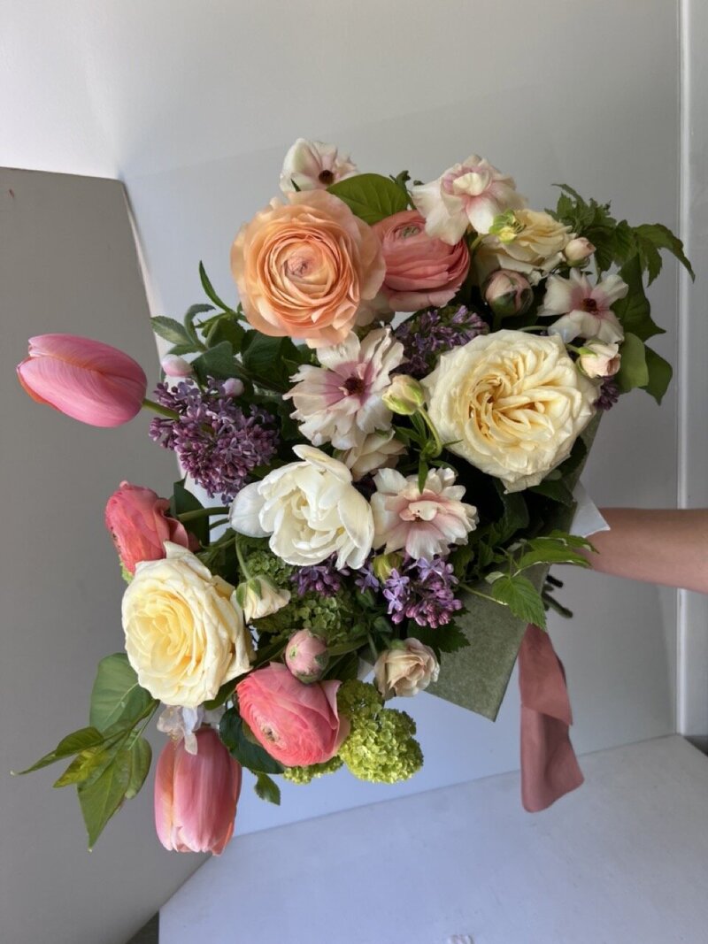 A large hand tied Mother's day bouquet is held up featuring tulips, ranunculus, roses and more. 