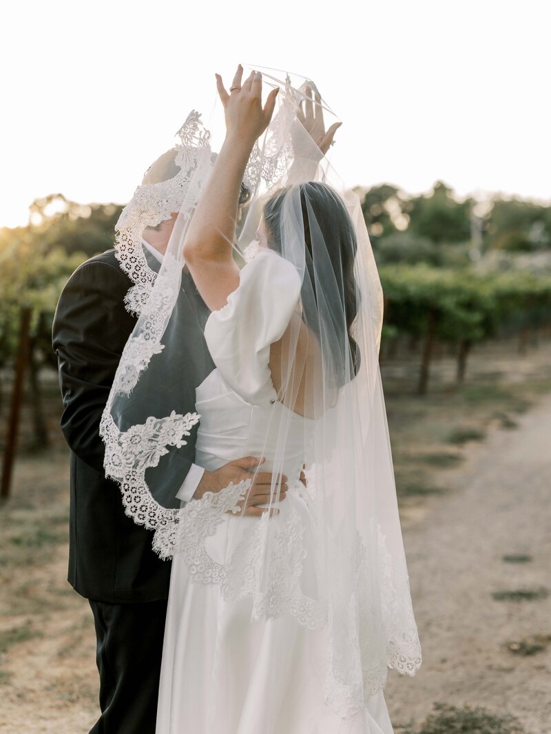  bride and Groom stand in the vineyard in San Luis Obispo during their wedding day at sunset, and she lifts the veil gently over his head and a documentary moment